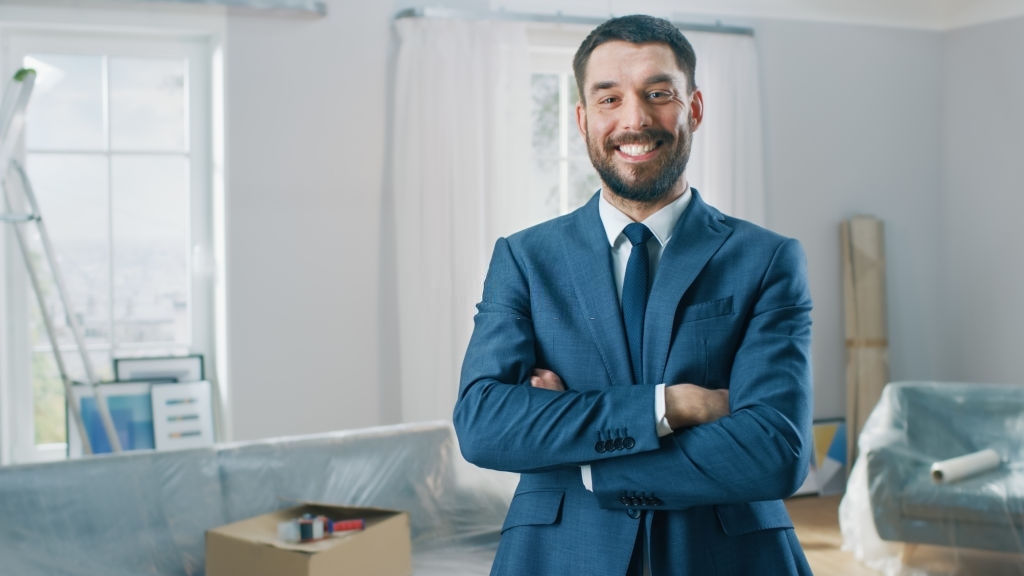 Successful Real Estate Agent in a Suit Smiles and Offers Keys From a New Apartment. Standing in the Middle of Room that Being Renovated. Spacious New House for Sale by Professional Real Estate Broker.